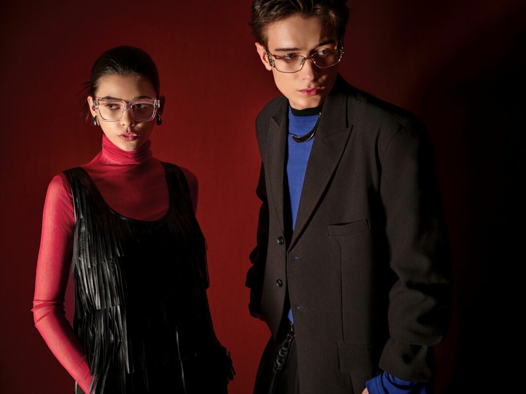Two models posing in stylish eyeglasses against red background.